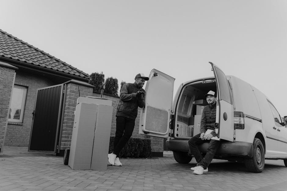 A man wearing a white t-shirt, light blue jeans, white sneakers, and a black cap is seen outside a residential property, carrying three cardboard boxes stacked together during a home relocation. The m