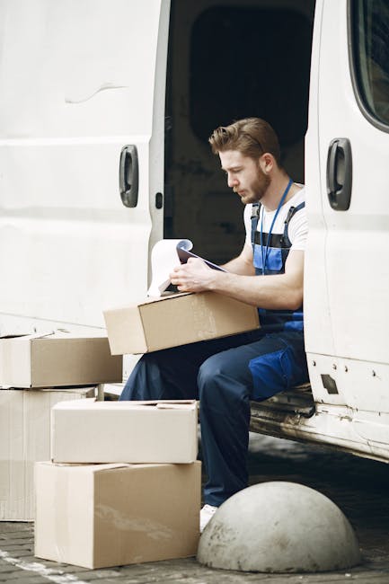 A man wearing a blue uniform with reflective stripes is sitting on the open sliding door of a white delivery van parked on a pavement, surrounded by cardboard boxes of various sizes. He is holding a c
