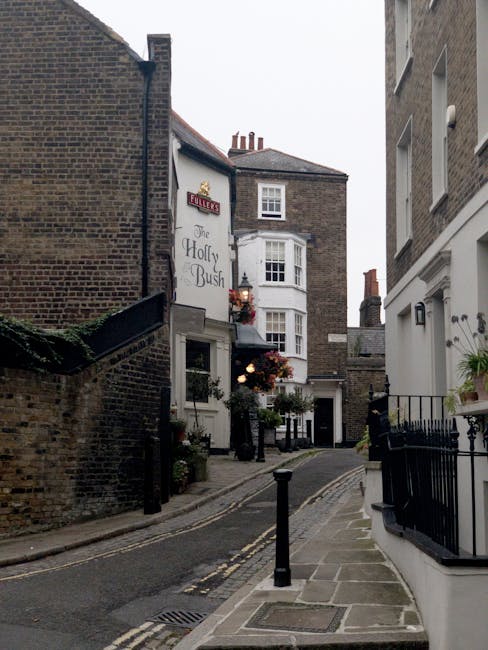 Two men are engaged in a home relocation process, lifting and loading cardboard boxes and a large flat-packed item into the back of a white van. One man, wearing a cap, glasses, and a padded jacket, i