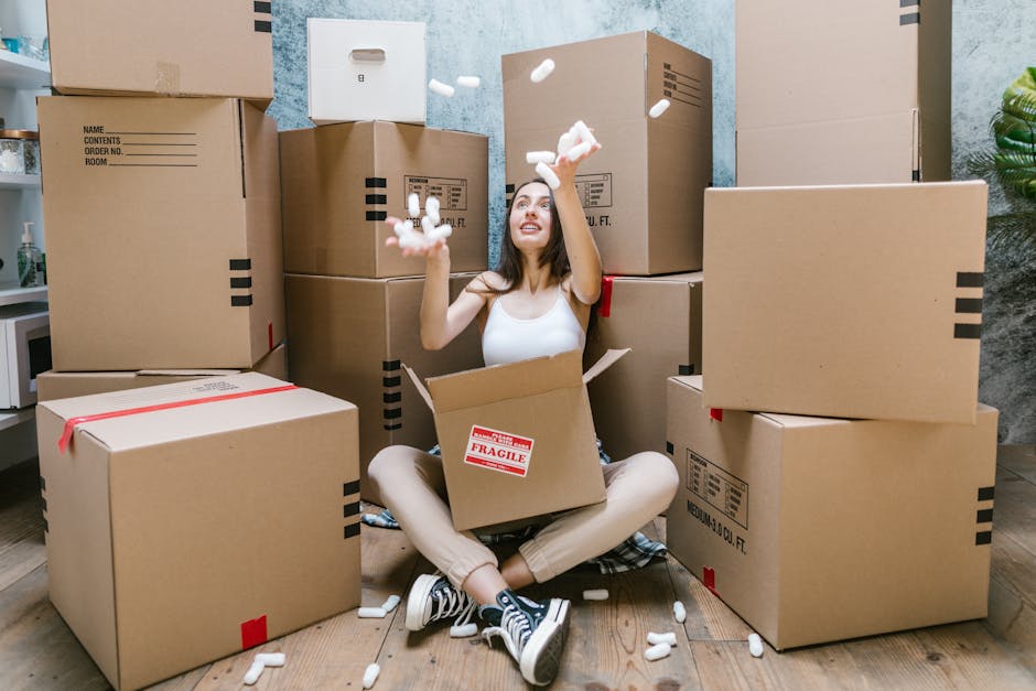 A young woman sitting on a wooden floor surrounded by large cardboard moving boxes inside a home, with more boxes stacked behind her. She is wearing a white tank top, beige pants, and black-and-white